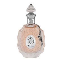 The image shows a decorative perfume bottle:  The bottle has an ornate, multi-faceted glass design with a textured surface and a pale pink tint, suggesting a delicate fragrance within. It features a clear glass stopper with an intricate cut pattern on top. A metallic silver chain with a charm and a small black stone is attached to the neck of the bottle. The front of the bottle displays a white oval label with Arabic calligraphy and the name "ROUAT AL MUSK" in Roman characters below.