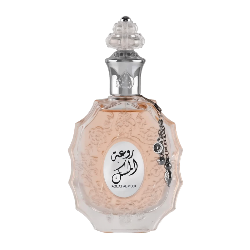 The image shows a decorative perfume bottle:  The bottle has an ornate, multi-faceted glass design with a textured surface and a pale pink tint, suggesting a delicate fragrance within. It features a clear glass stopper with an intricate cut pattern on top. A metallic silver chain with a charm and a small black stone is attached to the neck of the bottle. The front of the bottle displays a white oval label with Arabic calligraphy and the name "ROUAT AL MUSK" in Roman characters below.