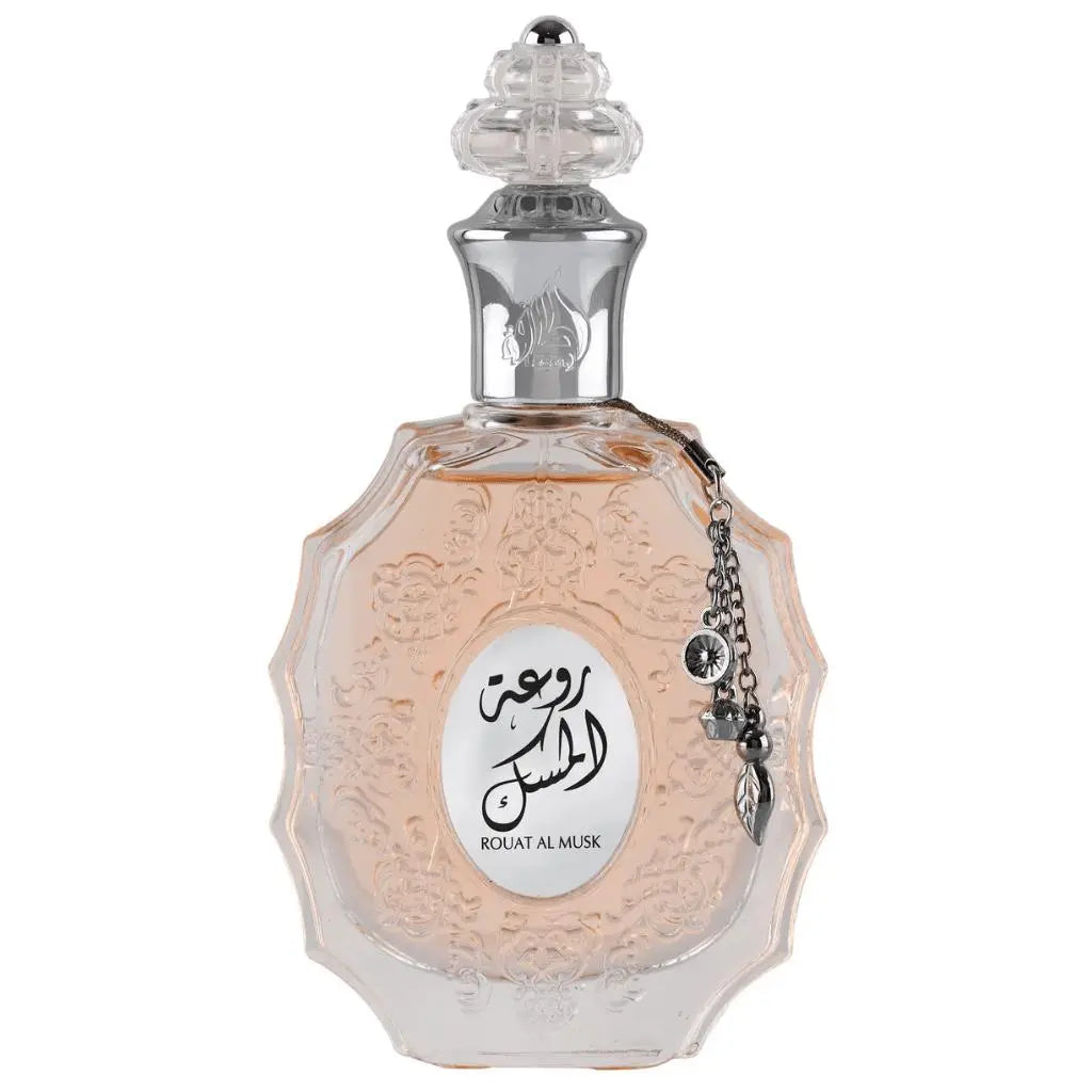 The image shows a decorative perfume bottle:  The bottle has an ornate, multi-faceted glass design with a textured surface and a pale pink tint, suggesting a delicate fragrance within. It features a clear glass stopper with an intricate cut pattern on top. A metallic silver chain with a charm and a small black stone is attached to the neck of the bottle. The front of the bottle displays a white oval label with Arabic calligraphy and the name "ROUAT AL MUSK" in Roman characters below.