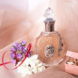 The image shows a perfume bottle on a soft, pastel background:  The clear glass bottle has a textured design and contains a pale pink perfume. An intricate glass stopper sits atop the bottle. A decorative metallic chain with a charm dangles from the neck of the bottle. The bottle label features Arabic calligraphy and the English inscription "ROUAT AL MUSK."