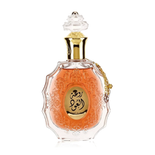 he image features an ornate perfume bottle:  The bottle is made of clear glass with a decorative texture and a central amber-colored panel. A clear and intricate glass stopper tops the bottle. A golden metallic chain with a charm is attached to the neck of the bottle. Arabic calligraphy is featured within the amber panel, and the name "ROUAT AL OUD" is inscribed in English below the calligraphy.