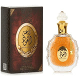 The image shows a perfume bottle and its box:  The perfume bottle is made of clear glass with an ornate design and features an amber-colored central area with decorative elements. Arabic calligraphy is displayed in the central area, with "ROUAT AL OUD" inscribed in English below it. The bottle has a clear, intricately designed stopper and a golden chain with a charm around its neck.