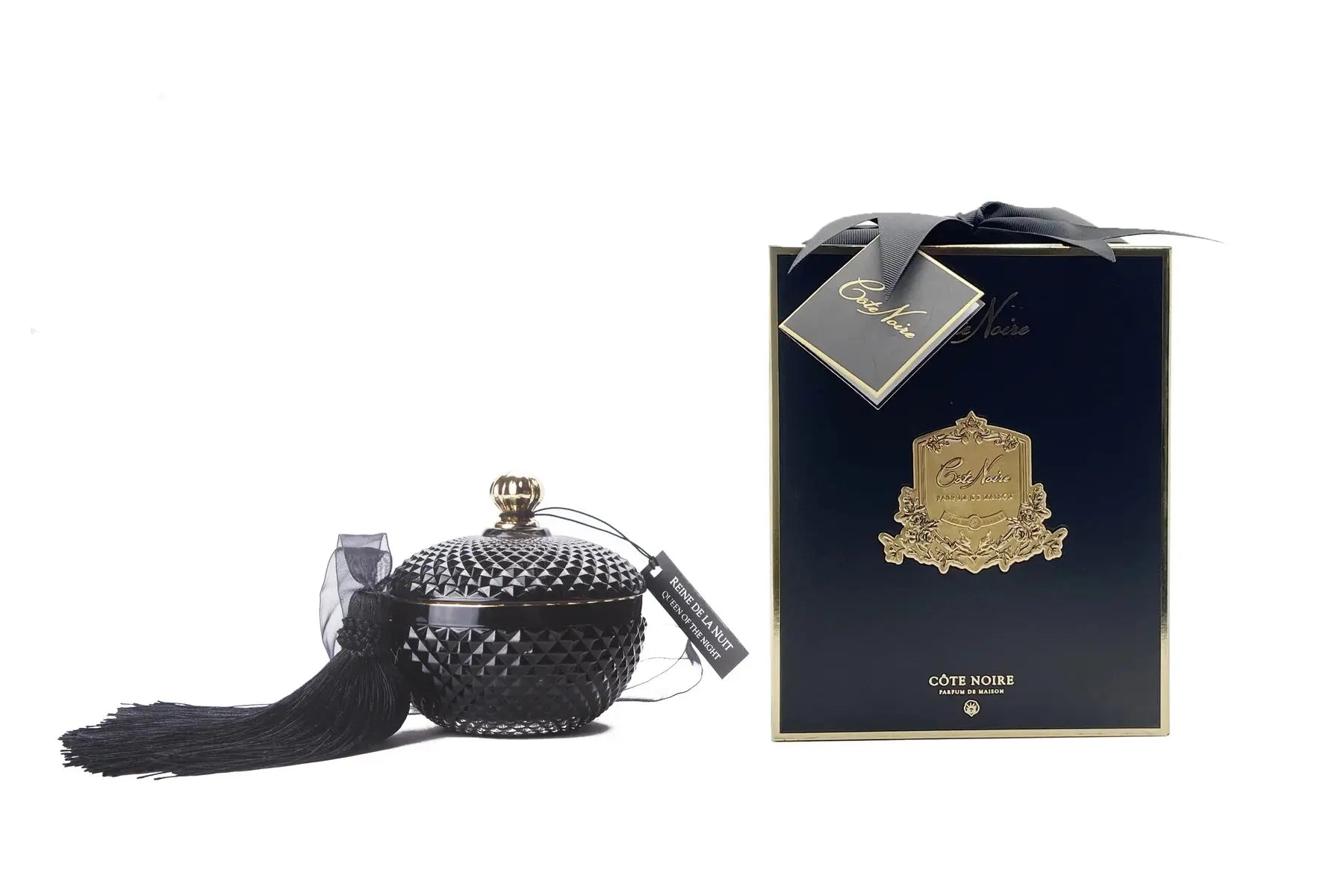 The image features a sophisticated product display with a candle and its accompanying packaging. On the left, there is a black, textured, round candle with a golden top and a long black tassel. Beside the candle is a transparent candle holder. On the right, there is an elegant dark blue box with golden accents and a black ribbon. The box and the candle have matching branding that includes intricate golden designs and text that enhances the luxurious feel of the product.