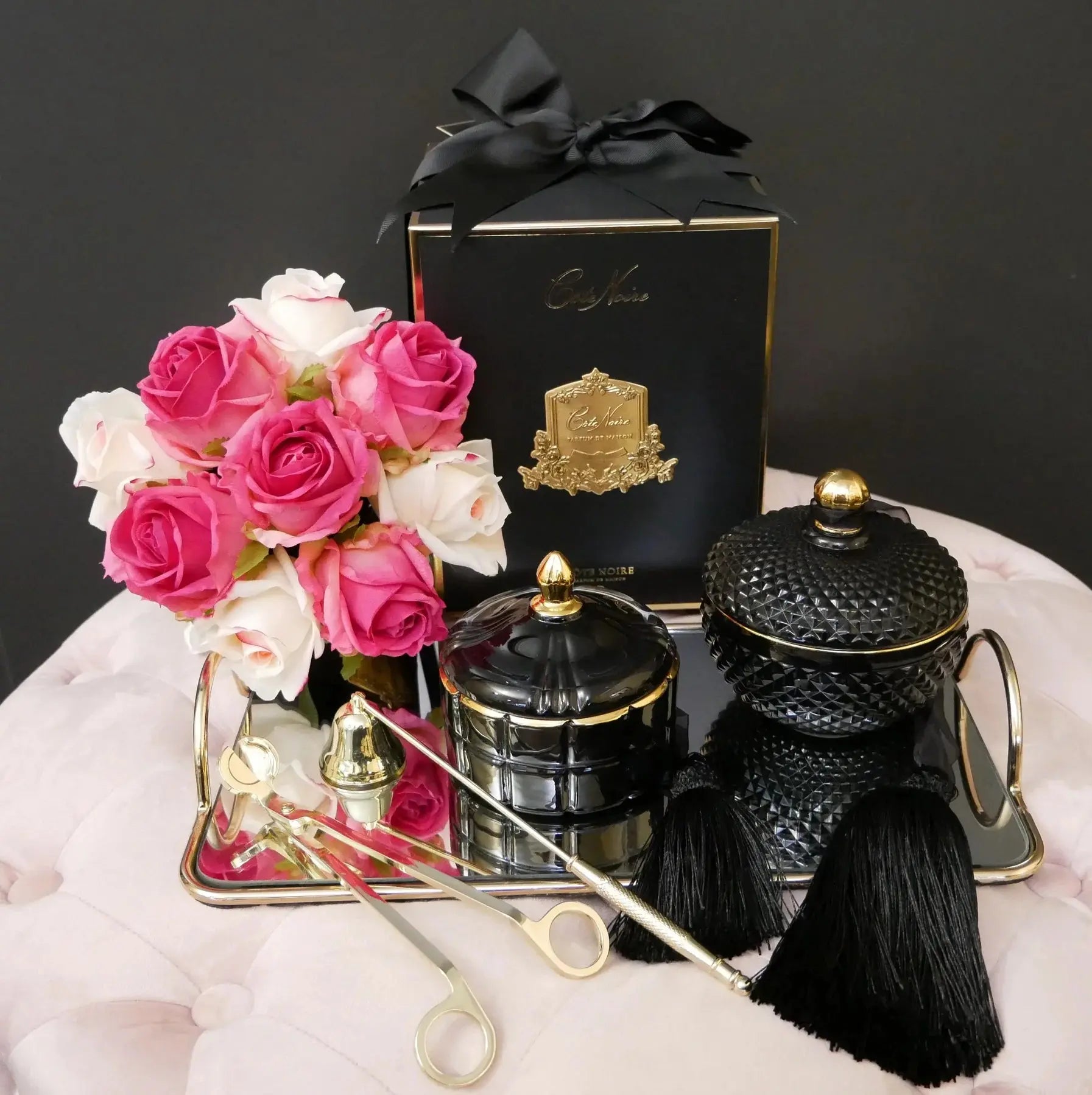 The image presents a beautifully arranged product display featuring luxury candles and decorative elements. Two round, textured candles with golden accents and long black tassels are displayed, one in black and the other in a dark charcoal. They are set on a mirrored tray beside a bouquet of vibrant pink and white roses held in a golden holder, adding a fresh and elegant touch to the arrangement. In the background, there is a dark blue box with a golden emblem and a black ribbon.