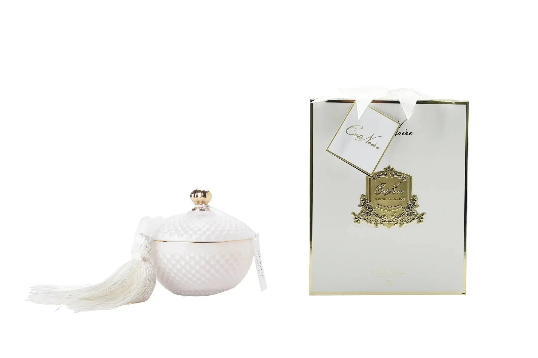 A round, Art Deco-style candle container in a white color with a textured diamond pattern, topped with a gold-colored knob and adorned with a matching white tassel. The container is accompanied by a white and gold rectangular gift box with a label that reads 'Côte Noire' in an elegant script. The box also features a gold emblem and a hanging tag.