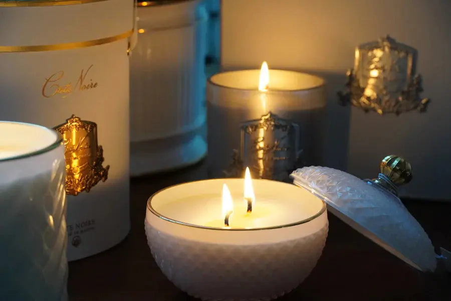 A cozy setting featuring several lit candles from Côte Noire. The focus is on a white, round, Art Deco-style candle container with a textured diamond pattern and a gold-colored knob, which is open with the lid placed beside it. The candle inside has three lit wicks. In the background, additional candles in white and gray containers, each adorned with an ornate gold label, are also lit, creating a warm and inviting atmosphere.