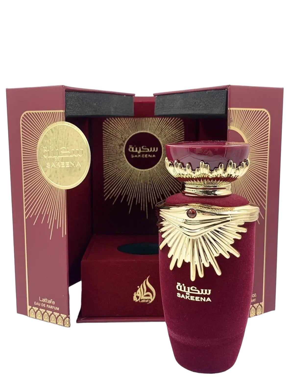 The image features an elaborate perfume presentation of "SAKEENA" by Lattafa. The box is a deep burgundy color with golden accents and a geometric, radial design emanating from a circular emblem that reads "SAKEENA" in Arabic script. Inside the box, the perfume bottle is revealed—a luxurious maroon vessel with a golden, crown-like lid embellished with a gemstone. The bottle rests on a velvet-like fabric, enhancing the feeling of opulence. On the front of the bottle, "SAKEENA" is written in Arabic.
