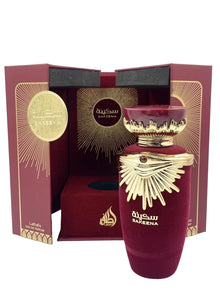 The image features an elaborate perfume presentation of "SAKEENA" by Lattafa. The box is a deep burgundy color with golden accents and a geometric, radial design emanating from a circular emblem that reads "SAKEENA" in Arabic script. Inside the box, the perfume bottle is revealed—a luxurious maroon vessel with a golden, crown-like lid embellished with a gemstone. The bottle rests on a velvet-like fabric, enhancing the feeling of opulence. On the front of the bottle, "SAKEENA" is written in Arabic.