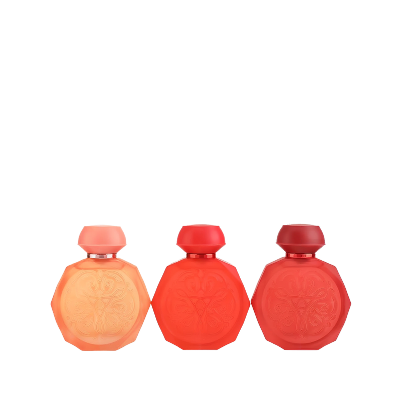 A set of three Gissah perfume bottles in a range of red hues, each with an octagonal shape and intricate embossed designs on the front. The bottles feature smooth, matching caps and come in shades of peach, vibrant red, and deep red. The elegant and minimalist design highlights the gradient of colors, making the set visually appealing and sophisticated.