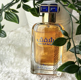 A square glass perfume bottle with a golden-yellow liquid is showcased. The bottle has a blue and white checkered cap and displays the name "SHAFAQ" in blue letters and Arabic script on its front. Below, in smaller print, is "By Ard al Zaafaran." The bottle is placed on a fluffy white textile surface, and there are green leaves in the upper right corner, possibly part of a houseplant, which adds a natural touch to the composition. The background is neutral and bright.