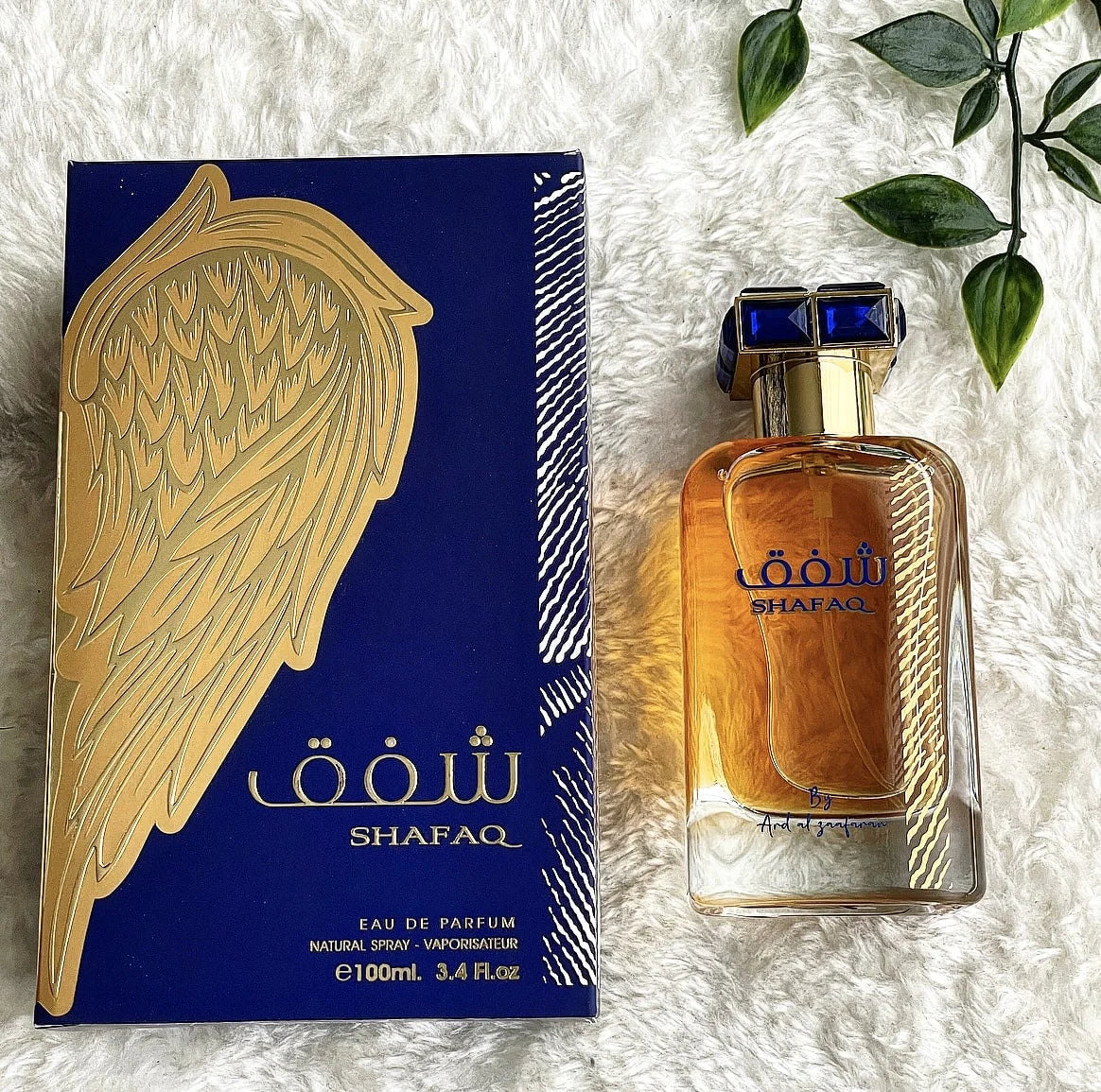 A square glass perfume bottle with a golden-yellow liquid is displayed next to its packaging. The bottle has a blue and white checkered cap and features the name "SHAFAQ" in blue letters with Arabic script. Below, in small print, is "By Ard al Zaafaran."  The bottle and box are placed on a fluffy white textile surface, with green leaves peeking into the frame from the top right corner.