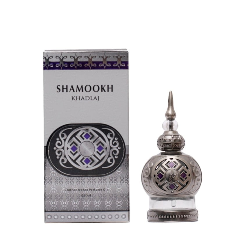 The image shows a product called "Shamookh" by Khadlaj, which is a concentrated perfume oil. The perfume comes in a 20ml spherical bottle with a silver finish, featuring intricate patterns and a pointed cap that adds a touch of elegance. The design includes decorative elements with purple accents, enhancing its luxurious appearance. The packaging box matches the bottle's design, with a silver and purple theme, featuring the product name "Shamookh" in bold letters and additional ornamental details.