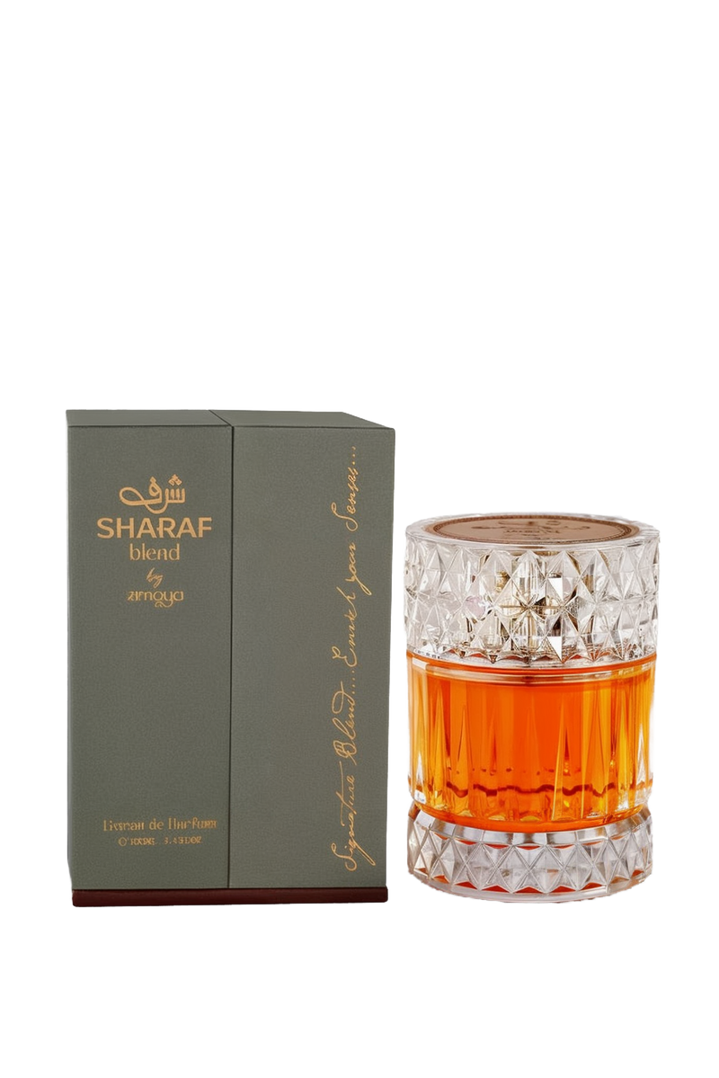 A cylindrical glass perfume bottle with a crystal-cut pattern at the top and bottom, filled with an amber-colored fragrance labeled “Sharaf Blend by Zimaya,” standing beside a dark green box embossed with gold Arabic and English text.
