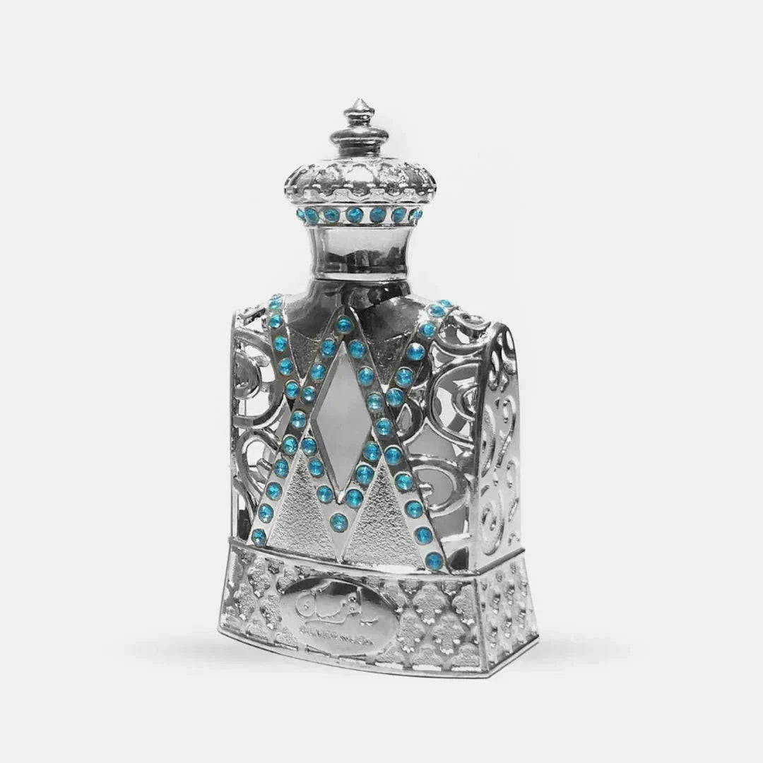 The image features an ornately designed perfume bottle, resembling a piece of classic royal attire or regalia. The body of the bottle is crafted in silver with elaborate openwork filigree details that lend it a lace-like appearance. Adorned with a multitude of bright blue gemstones, they are strategically placed in V-shaped patterns across the front of the bottle, adding a striking contrast to the metallic silver. The neck of the bottle, also silver, is encrusted with a band of these blue gemstones.