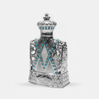 The image features an ornately designed perfume bottle, resembling a piece of classic royal attire or regalia. The body of the bottle is crafted in silver with elaborate openwork filigree details that lend it a lace-like appearance. Adorned with a multitude of bright blue gemstones, they are strategically placed in V-shaped patterns across the front of the bottle, adding a striking contrast to the metallic silver. The neck of the bottle, also silver, is encrusted with a band of these blue gemstones.