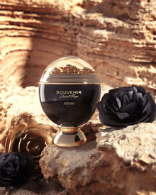 The image depicts an elegant perfume bottle placed on a rocky desert-like terrain, resonating with the name "SOUVENIR Desert Rose AFNAN" inscribed in gold on the bottle's black surface. The bottle has a rounded base and is topped with a clear dome that encases golden flakes, giving the impression of a miniature desert landscape.