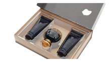 The image shows an elegant gift set by Afnan Perfumes from their "SOUVENIR Desert Rose" collection. The set is displayed in a beige presentation box with a clear lid, revealing its contents. Inside, there is a dark, glossy perfume bottle with a spherical design and a golden cap, positioned centrally. On either side of the perfume bottle are two matching dark tubes.  Both tubes and the perfume bottle feature the "SOUVENIR Desert Rose" branding in elegant gold lettering