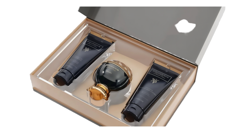 The image shows an elegant gift set by Afnan Perfumes from their "SOUVENIR Desert Rose" collection. The set is displayed in a beige presentation box with a clear lid, revealing its contents. Inside, there is a dark, glossy perfume bottle with a spherical design and a golden cap, positioned centrally. On either side of the perfume bottle are two matching dark tubes.  Both tubes and the perfume bottle feature the "SOUVENIR Desert Rose" branding in elegant gold lettering