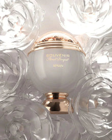 The image shows a perfume bottle with a clean and elegant design. The bottle has a spherical white body with a golden band that reads "SOUVENIR Floral Bouquet AFNAN." Above the band is a clear dome, encasing what appears to be a cluster of golden petals or flakes, giving a sense of luxury. The bottle rests on a golden pedestal base that complements the golden accents.