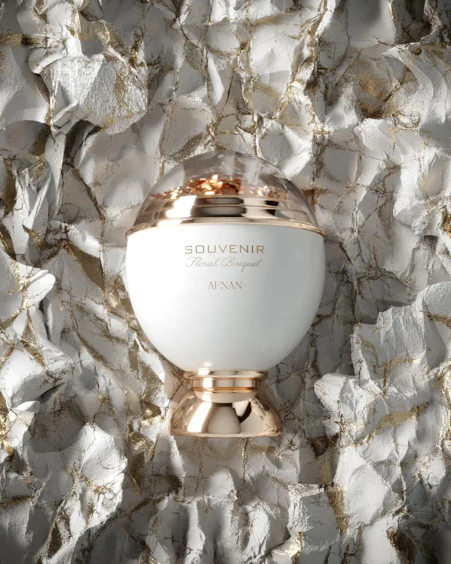 The bottle, named "SOUVENIR Floral Bouquet AFNAN," has a spherical white body with a reflective rose gold band around its center. The top of the bottle features a clear dome that encases what appears to be gold flakes or petals, adding to the luxurious feel of the product. The bottle's base is also rose gold, complementing the band and the warm tones in the background. 