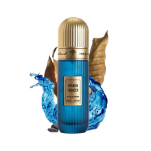 An image featuring a cylindrical perfume bottle with a striking blue color and vertical ribbed texture. The bottle has a luxurious gold cap and a label stating "SPANISH TOBACCO" in bold, with "Extrait de parfum 100 ml 3.4 fl.oz." underneath. Accompanying the bottle are visual elements like a dried brown tobacco leaf, an ornate golden-brown container, and vibrant blue ink or liquid splashing dynamically around the bottle. 
