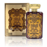 The image depicts a luxurious perfume set that includes a bottle and its packaging. The perfume bottle has a deep burgundy color with a gold label that features elaborate scrollwork and Arabic calligraphy reading "AL IBDAA." The same design and text appear on the gold-colored box behind the bottle, which also states "EAU DE PARFUM NATURAL SPRAY 100ml 80% Vol 3.4 FL.OZ" in smaller English text.  he packaging also indicates that the fragrance is for women.