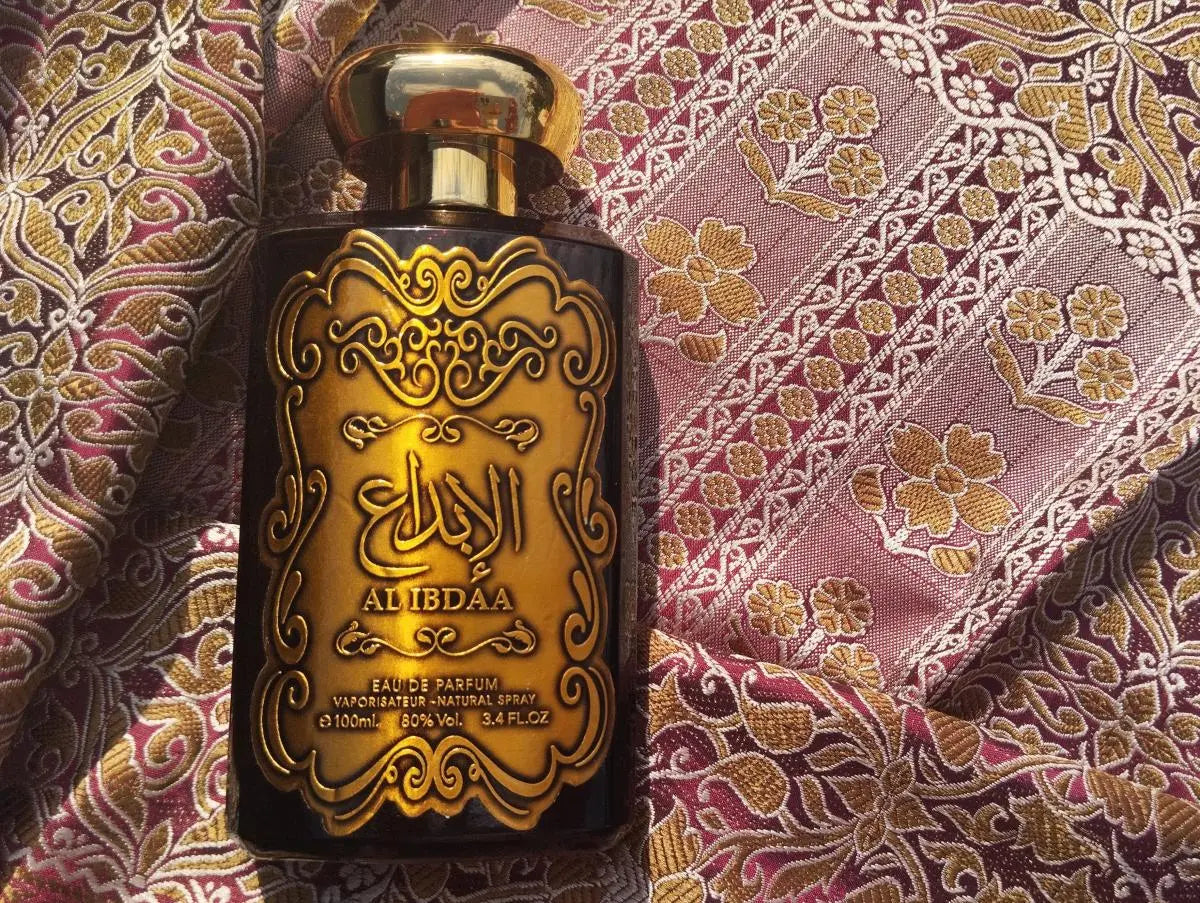 The image shows a perfume bottle with a black body and a golden label featuring intricate scrollwork and Arabic calligraphy that reads "AL IBDAA".  The bottle is placed against a richly patterned fabric with a floral and paisley design in hues of purple, pink, and gold, suggesting an exotic and luxurious setting that matches the opulence of the perfume bottle.
