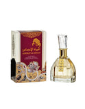 The image shows a fragrance product called 'Ameerat Al Ehsaas' by 'Ard Al Zaafaran'. The set includes a perfume bottle and its corresponding packaging box. The box has a maroon top with ornate gold patterns and Arabic calligraphy, and a complex paisley and floral design in gold, black, and maroon on the lower half. The bottle is transparent with a golden-yellow perfume inside and a rectangular gold label with a row of rhinestones. It has a gold neck and a clear, faceted cap. 