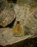 The image features a perfume bottle named 'Ameerat Al Ehsaas' positioned on a rocky surface, likely outdoors. The bottle is made of clear glass with a golden-yellow tinted perfume inside. The design is elegant, with a broad base that narrows towards the top and a decorative golden band around the neck featuring a rectangular area filled with small gold-colored studs. It has a faceted clear glass cap that reflects the light, adding to the luxurious look of the product. 
