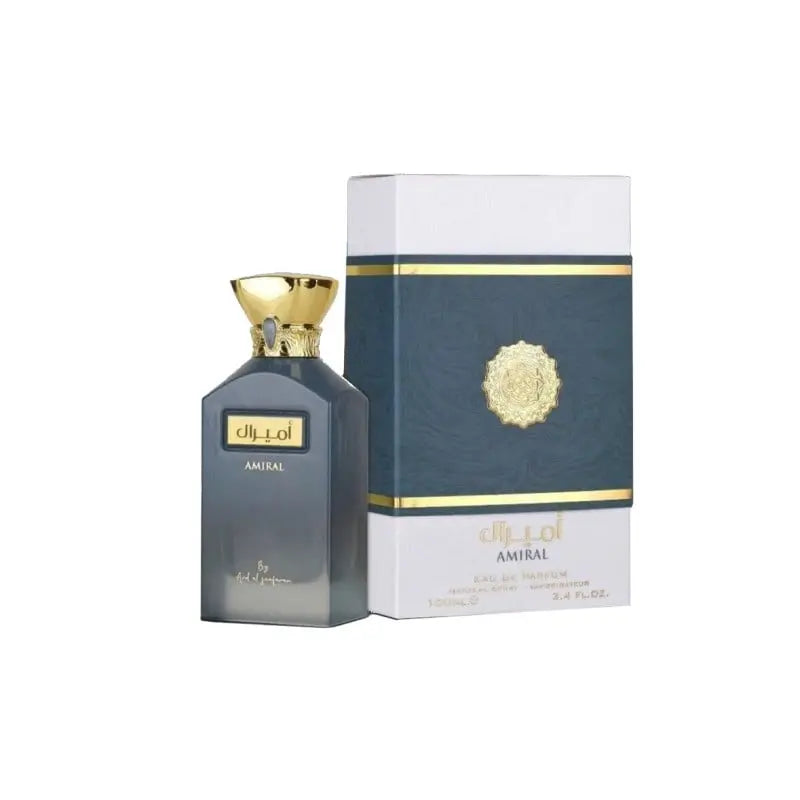 The image shows a bottle of AMIRAL perfume by Ard Al Zaafaran, next to its packaging. The bottle is matte grey with a gold cap designed like a crown, with a gold ring and a gemstone at the neck. The front of the bottle has the name "AMIRAL" in black on a gold plaque with Arabic script above it and "By Ard Al Zaafaran" written below. The packaging is a white box with a central dark blue panel bordered by gold lines. 