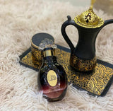 The image displays a bottle of Aroosat Al Emarat perfume on a patterned black and gold coaster. The perfume bottle has a dark, spherical body with a pink gradient at the bottom and a gold label with Arabic calligraphy in the center. It is topped with a clear, flower-shaped cap and a gold neck. Next to the perfume is a black traditional Arabian-style jug with a gold-patterned trim and a gold lid with intricate designs.