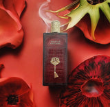 The image depicts a rectangular, deep red perfume bottle with a textured gold cap, set against a backdrop of vivid red flower petals and one prominent green and red flower.  The scene is artfully arranged to evoke a sense of opulence and exotic allure, with the red tones of the flowers complementing the bottle's design and suggesting a floral or rich scent.