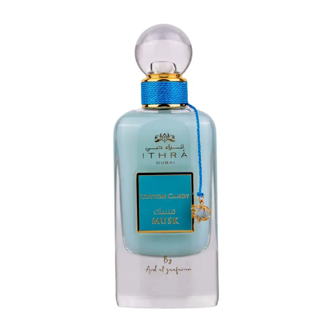 The image shows a perfume bottle with a clear, pale blue liquid. The bottle has a rounded rectangular shape with a clear spherical cap. Around the neck of the bottle is a blue ribbon, and there's a small gold charm attached, featuring what appears to be an animal or a crest. The front of the bottle has a teal label with gold trim and the words "COTTON CANDY MUSK" in capital letters. Above the label in smaller text is written "ITHRA DUBAI", and below it is "By Ard Al Zaafaran". 