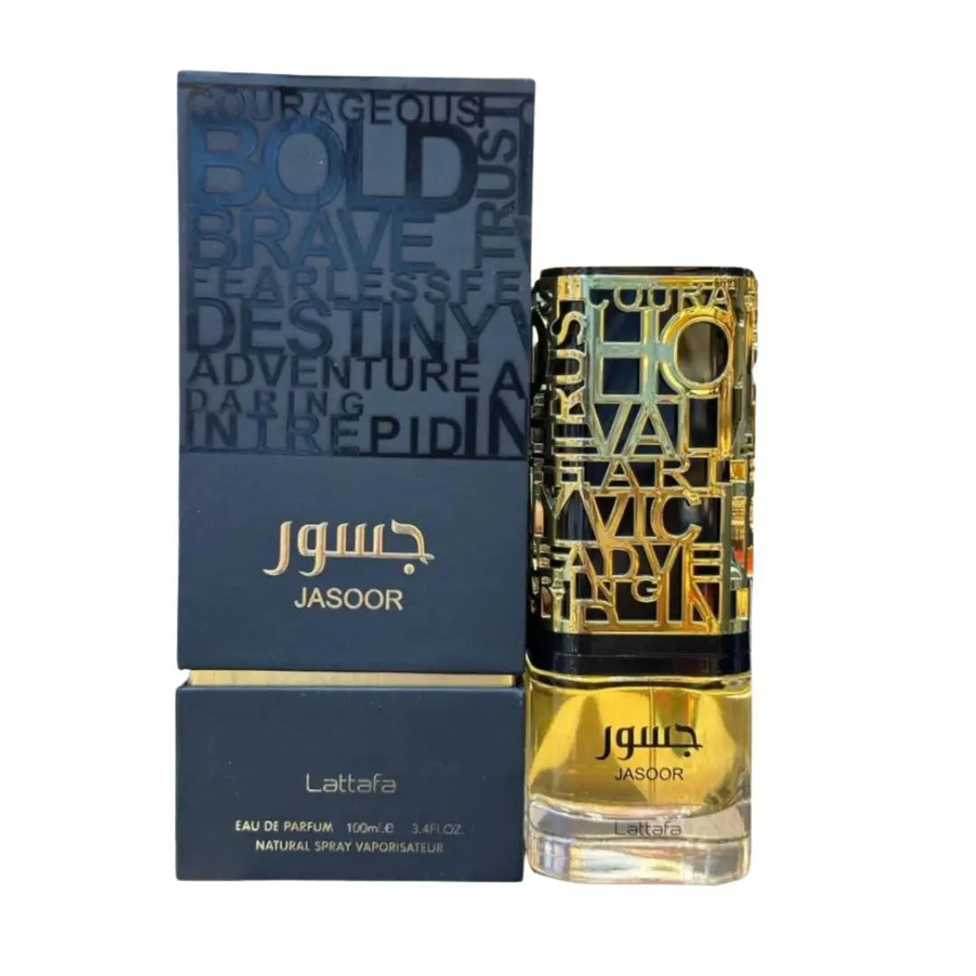 The image features a bottle of "JASOOR" perfume by Lattafa next to its packaging. The perfume bottle has a clear base with a yellow-amber colored fragrance, a black band near the top, and a gold-colored metallic overlay with cut-out letters. The packaging box is navy blue with a similar design to the bottle, featuring words like "BOLD", "BRAVE", and "DESTINY" in a lighter shade of blue. 
