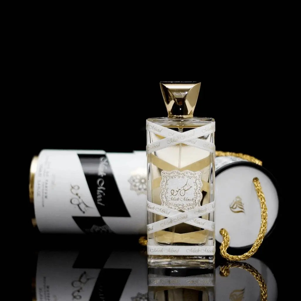 The image shows a luxurious perfume bottle with a clear body and golden-yellow liquid, paired with its packaging. The bottle features a white ribbon with the text "Musk Mood" printed on it, and a gold, ornate label in the center. The cap is a polished gold geometric shape. In the background, the cylindrical packaging is partially visible, decorated with black and white stripes and gold accents. A gold rope handle adds a decorative touch to the packaging.