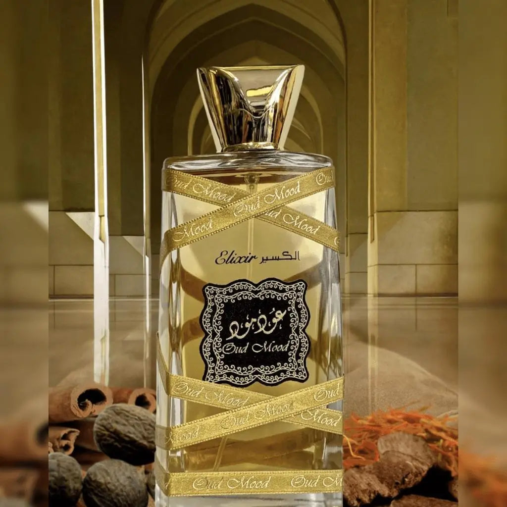 The image is of a perfume bottle set against an ornate background:  The perfume bottle is transparent with a golden-colored fragrance liquid inside. It has a gold cap in the shape of an inverted pyramid. Around the bottle is a golden ribbon with the words "Oud Mood" printed in black. The label on the bottle features intricate black designs and Arabic calligraphy, along with the word "Elixir" in English.