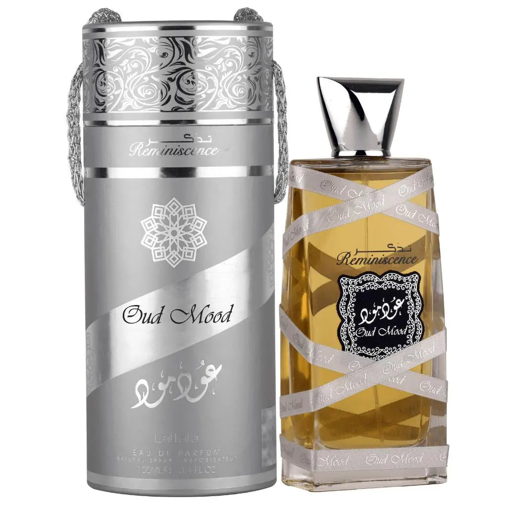 The image shows a perfume bottle next to its metallic container:  The perfume bottle is rectangular, made of clear glass with a silver ribbon wrapped around it, and filled with a golden-colored liquid. The ribbon has "Oud Mood" printed on it, and there's a central label with a black background and ornate white border featuring Arabic calligraphy and the word "Reminiscence" in English script. The bottle cap is silver and shaped like an inverted pyramid.