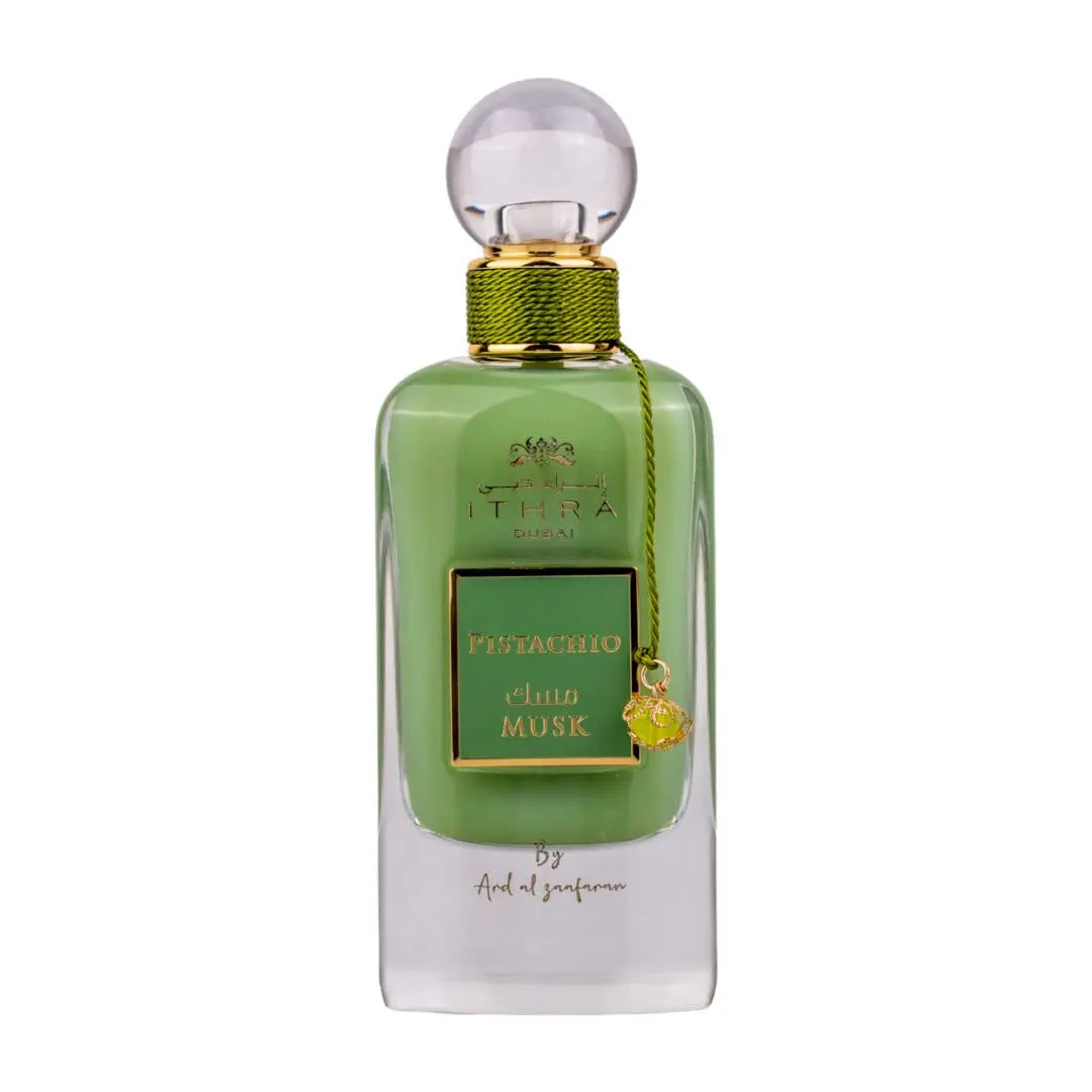 A bottle of Ithra Dubai Pistachio Musk perfume by Ard Al Zaafaran. The bottle has a clear glass design with a white gradient base, featuring a round glass cap. The liquid inside appears to be green, and there's a gold-colored brand emblem at the top, with the name of the scent on a green and gold label in the center. A small gold charm is attached to the neck of the bottle with a green string.