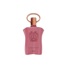 The image shows a pink perfume bottle with a metallic gold cap and a ring-shaped detail on top. The front of the bottle displays embossed text and a decorative emblem featuring the initials 'AP' in a stylized design, surrounded by a wreath-like motif. Below the emblem, the words "SUPREMACY GALA AFNAN" are printed in a simple, elegant font. The bottle has a smooth, matte finish, and there are small, pink gemstone-like embellishments around the neck.