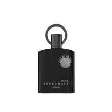The image shows a sleek, black perfume bottle with a modern design. The bottle has a cylindrical shape with a matte finish and features a metallic emblem in the center. Below the emblem, the text "NOIR SUPREMACY AFNAN" is printed in white, elegant font. The cap of the bottle is integrated into the design, creating a seamless look with a ring-shaped structure at the top. The overall aesthetic is luxurious and contemporary.