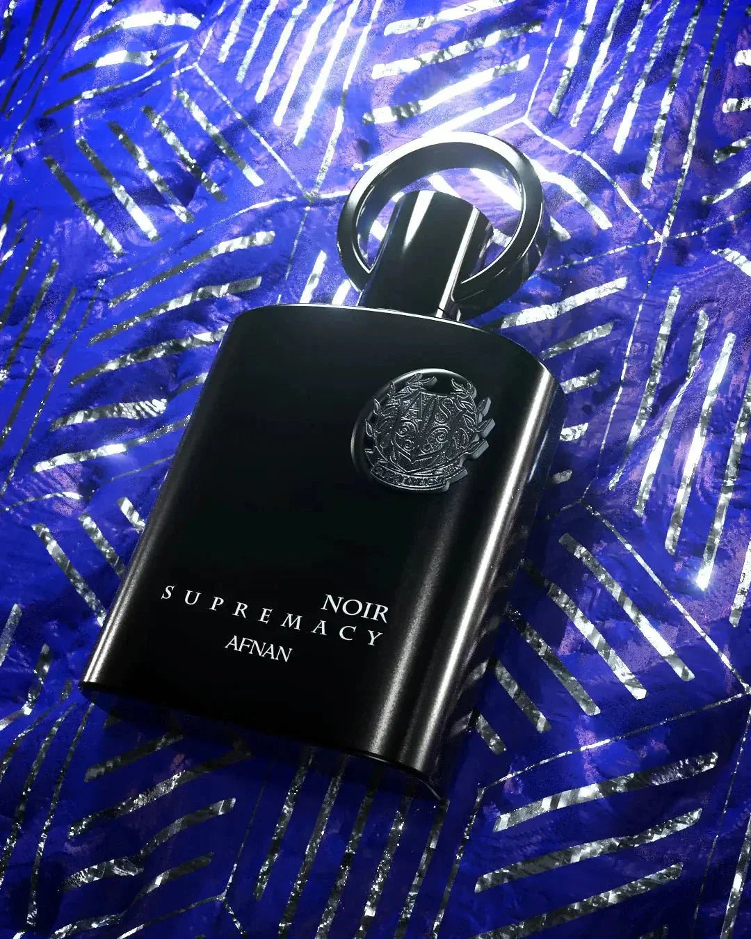 The image features an elegant black perfume bottle with a reflective, ring-shaped cap. The bottle is emblazoned with a silver emblem and the text "NOIR SUPREMACY AFNAN" in a sophisticated white font. It is placed against a vivid blue background adorned with a pattern of shiny, tropical palm leaves. The contrast between the dark bottle and the bright, luminescent blue creates a striking visual presentation, highlighting the product's luxurious appeal.