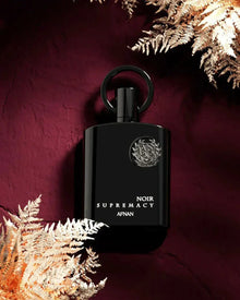 The image showcases a black perfume bottle with a silver emblem and white lettering that reads "NOIR SUPREMACY AFNAN". The bottle features a stylish ring-shaped cap and is set against a rich, burgundy background with golden-hued fern leaves partially framing the bottle. The luxurious color palette and the metallic sheen of the fern leaves give the image a warm, opulent feel, accentuating the exclusivity of the perfume.