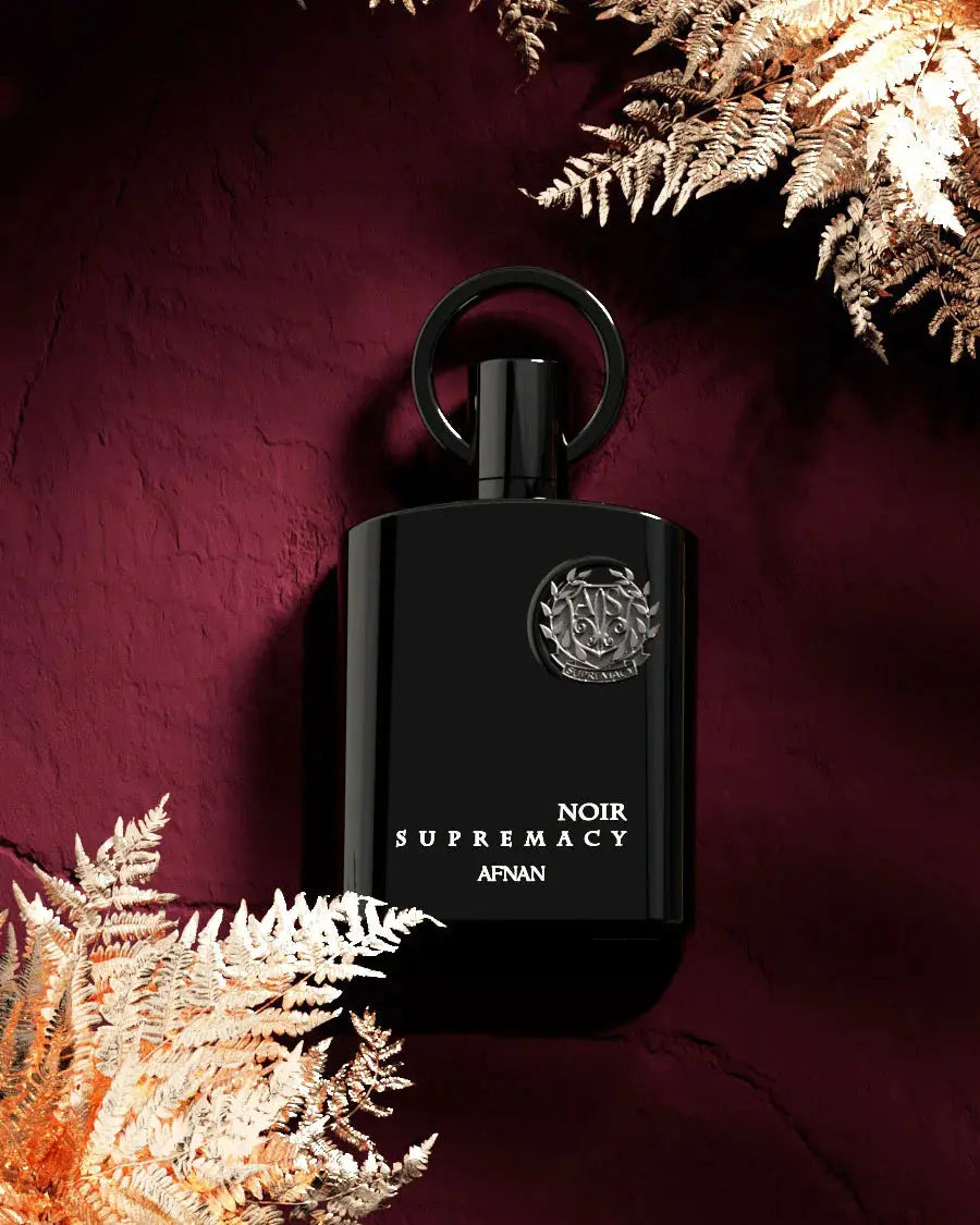 The image showcases a black perfume bottle with a silver emblem and white lettering that reads "NOIR SUPREMACY AFNAN". The bottle features a stylish ring-shaped cap and is set against a rich, burgundy background with golden-hued fern leaves partially framing the bottle. The luxurious color palette and the metallic sheen of the fern leaves give the image a warm, opulent feel, accentuating the exclusivity of the perfume.