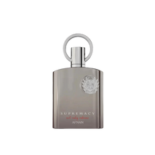The image shows a perfume bottle with a sleek, modern design. The bottle is a muted silver-gray with a matte finish and a flat, rectangular shape. The cap is silver and features a prominent circular loop, which adds a touch of sophistication. Emblazoned on the front of the bottle is a raised silver emblem that adds a 3D effect, and below it, the words "SUPREMACY not only intense AFNAN" are printed in a subtle, elegant font. 