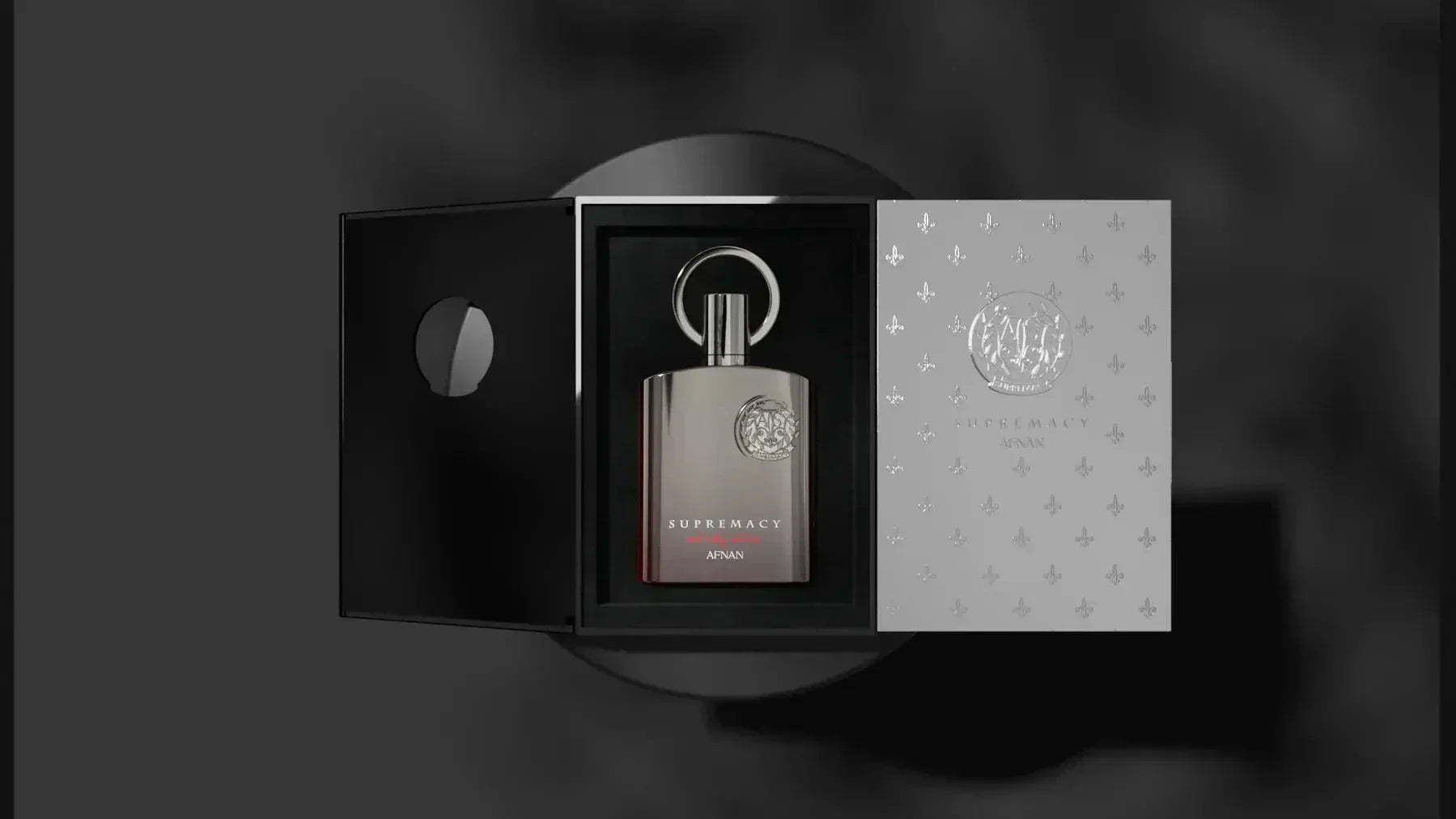 The image presents a metallic grey perfume bottle with a silver emblem, placed inside an open, elegant packaging. The left side of the packaging is black with a circular cutout revealing the bottle, while the right side has a silver finish with the bottle's emblem pattern repeated throughout. The front of the perfume bottle displays the text "SUPREMACY AFNAN" in white. The image is set against a monochromatic blurred background, highlighting the product's sophisticated design. 