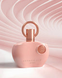 The image features a pink perfume bottle with a glossy finish, set against a textured background with a wavy, fabric-like pink design that creates a sense of motion. The bottle has a unique shape with rounded edges and a flat profile. It is adorned with a rose gold-colored ring-shaped cap and matching spray nozzle. The lighting highlights the bottle's curves and the emblem's detail. 