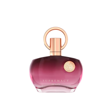 The image shows a perfume bottle with a distinctive design. It has a round, flat base with a gradient color that shifts from transparent to a deep pink hue towards the bottom. The cap is a metallic rose gold color, featuring a simple yet elegant circular design that loops around the nozzle. The front of the bottle has a metallic emblem with intricate detailing, adding a touch of luxury. The text on the bottle reads "SUPREMACY AENAN". 