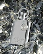 The image displays a matte silver perfume bottle of "SUPREMACY" by AFNAN positioned amidst a bed of crystal clear glass stones. The bottle has a clean, rectangular design with rounded edges and features a metallic silver cap with a loop design. On its surface, there is an embossed emblem that adds a touch of luxury to the design, and the brand name "AFNAN" is printed below. 