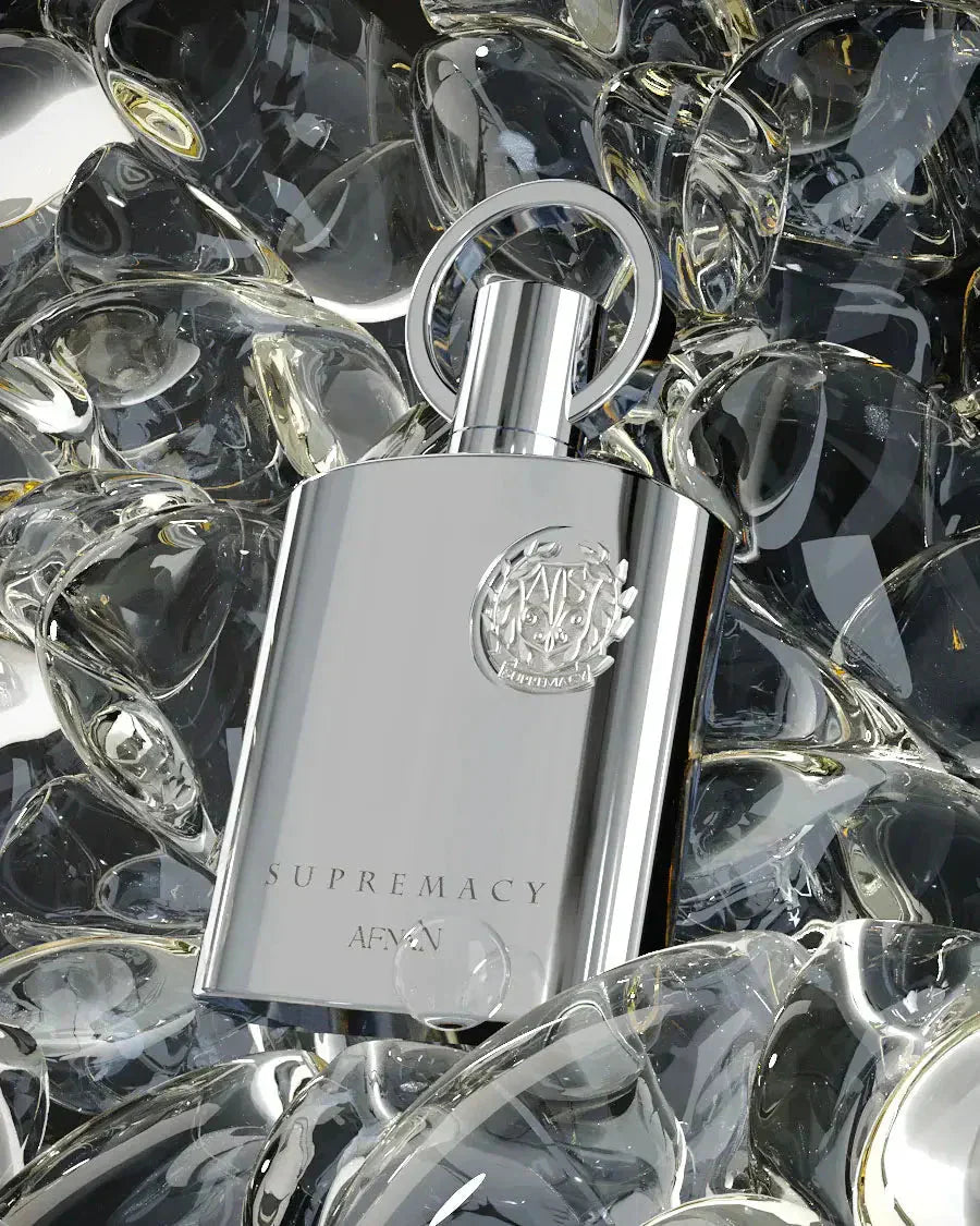 The image displays a matte silver perfume bottle of "SUPREMACY" by AFNAN positioned amidst a bed of crystal clear glass stones. The bottle has a clean, rectangular design with rounded edges and features a metallic silver cap with a loop design. On its surface, there is an embossed emblem that adds a touch of luxury to the design, and the brand name "AFNAN" is printed below. 