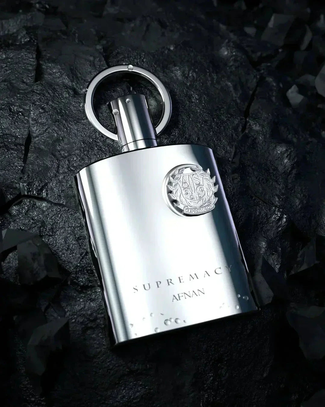 The image shows a silver perfume bottle from the "SUPREMACY" line by AFNAN. The bottle is placed on a textured, dark stone background that contrasts with its shiny metallic finish. It has a modern, sleek design with a reflective surface, and is accented with a circular metallic loop atop its cap. An intricate emblem is embossed on the bottle, adding to the luxurious look.   Water droplets are visible on the bottle, suggesting a fresh appeal and highlighting the bottle's metallic sheen. 